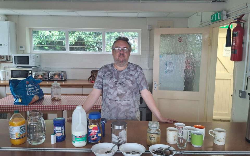 A man stood behind a kitchen counter with tea, coffee and sugar in front of him.