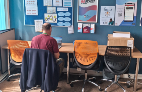 A man sat at a desk  on a computer with posters on the wall in front of him. 