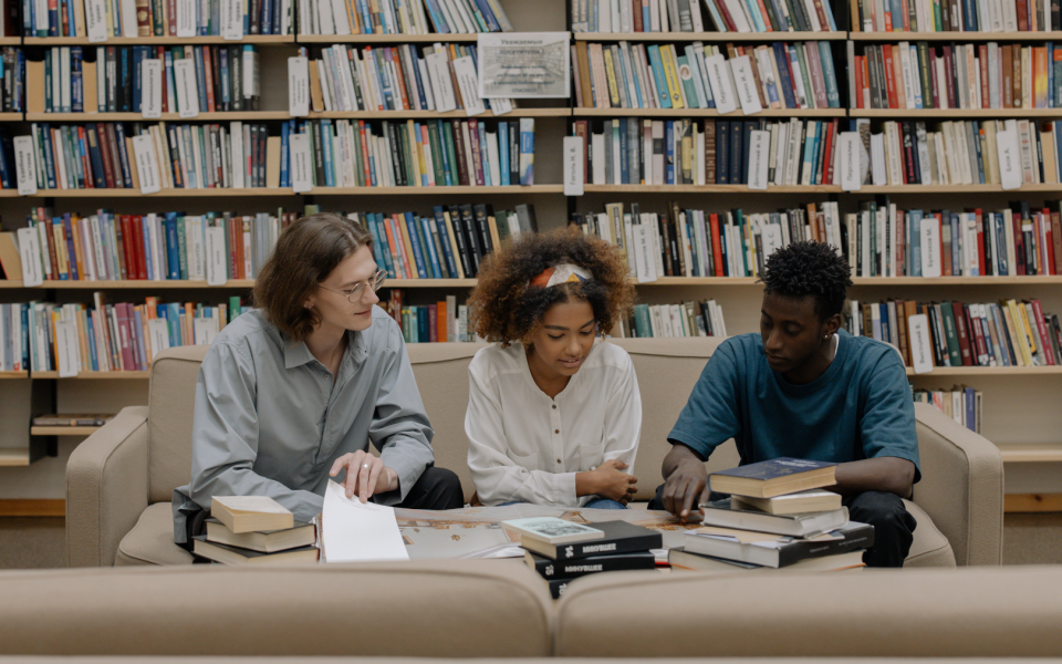 3 students sitting on a sofa in a library looking at books together