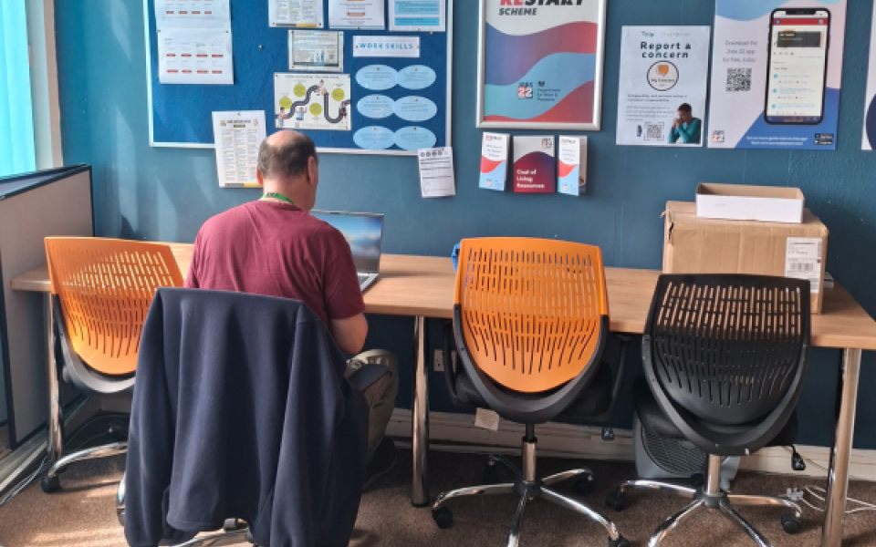 A man sat at a desk  on a computer with posters on the wall in front of him. 